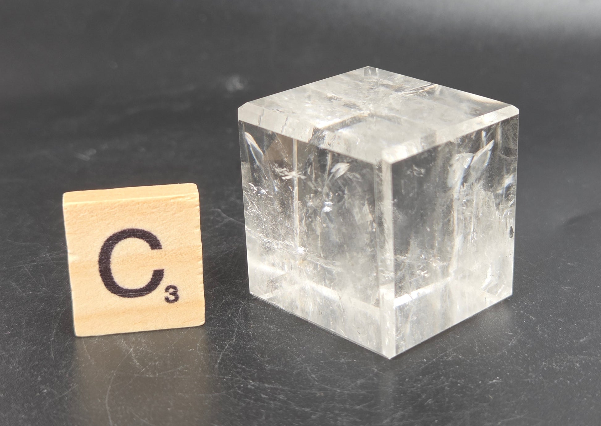 A clear, faceted crystal cube with natural inclusions sits on a dark gray surface next to a wooden Scrabble tile marked 'C3' for size comparison.