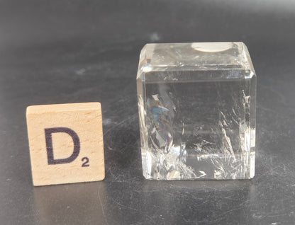 A small clear glass cube with visible scratches and reflections, placed on a dark gray surface next to a wooden Scrabble tile showing the letter D.