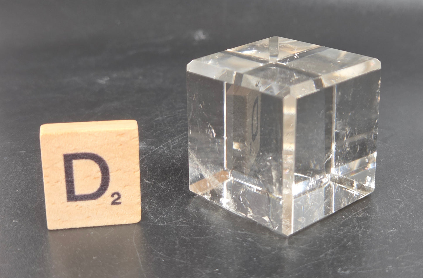 A clear glass cube next to a wooden Scrabble tile with the letter D and subscript 2, set on a dark gray surface.