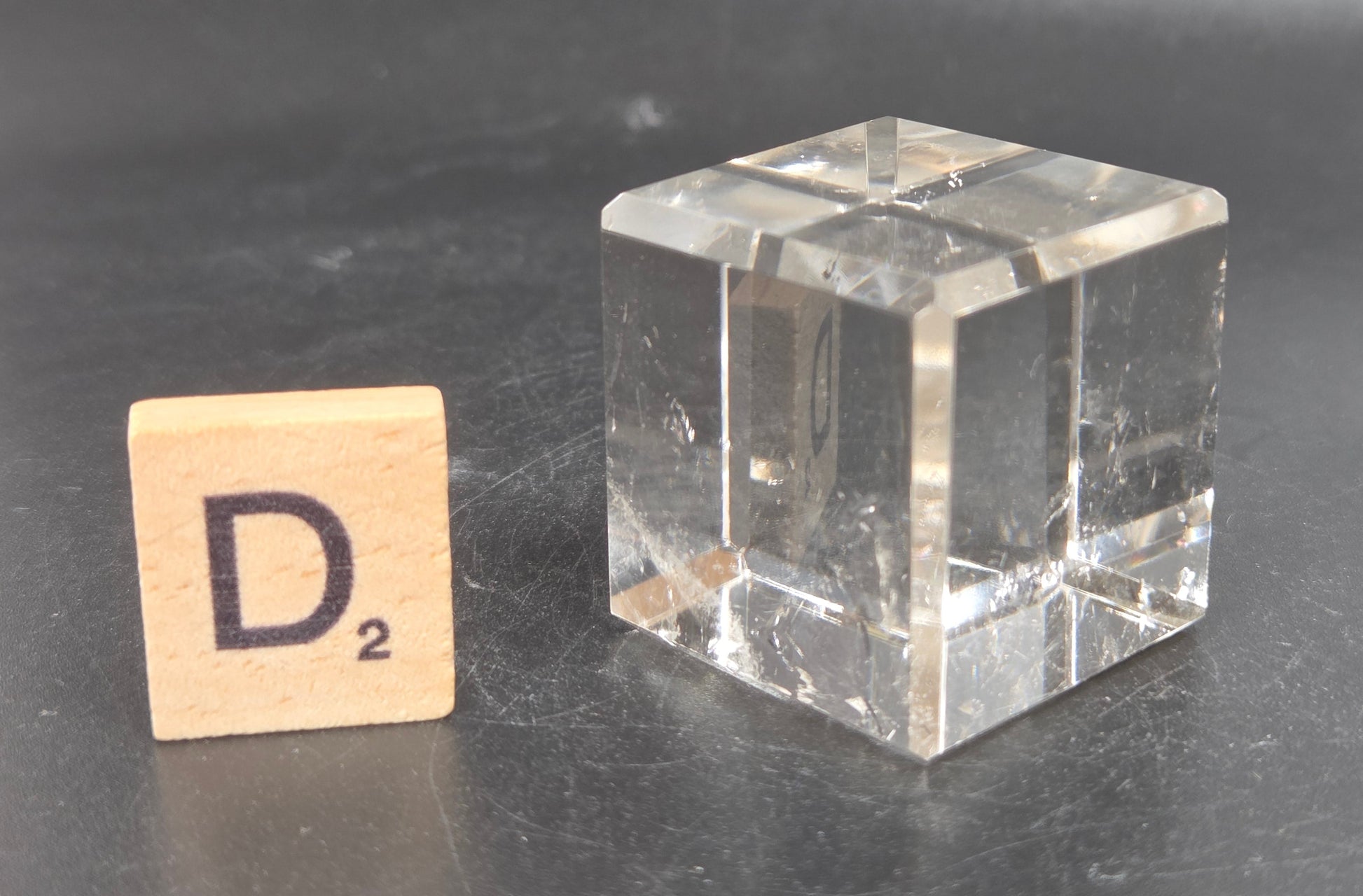 A clear glass cube next to a wooden Scrabble tile with the letter D and subscript 2, set on a dark gray surface.