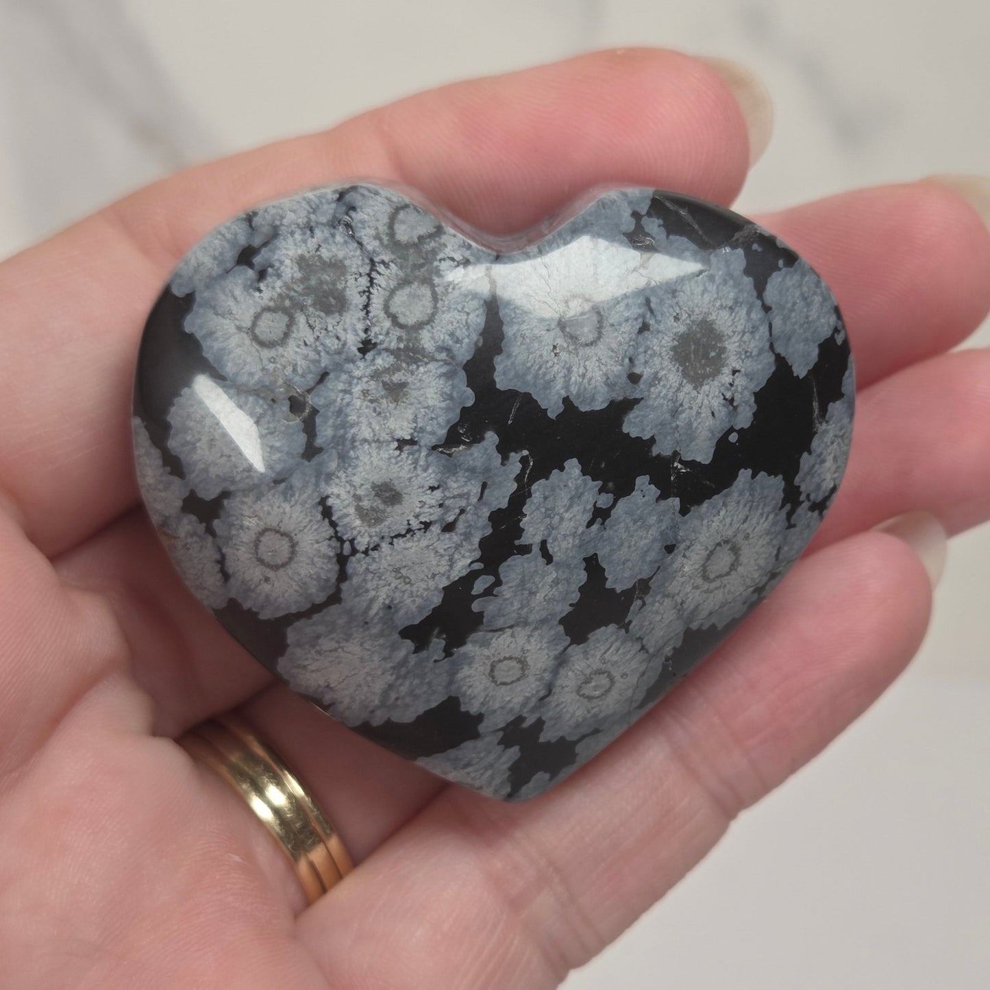 A heart-shaped polished stone with a black and gray floral or coral-like pattern, held in a person's hand against a light marble background.