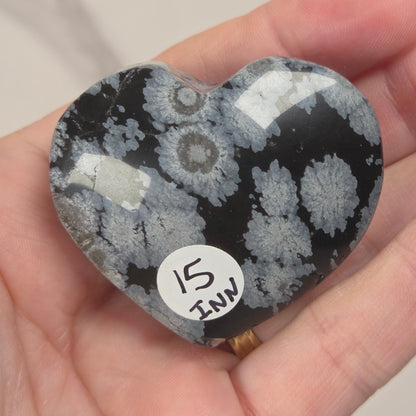 A heart-shaped polished stone with a black and white agate-like pattern, held in a person's hand. A small white sticker on the stone reads '15 INK'.