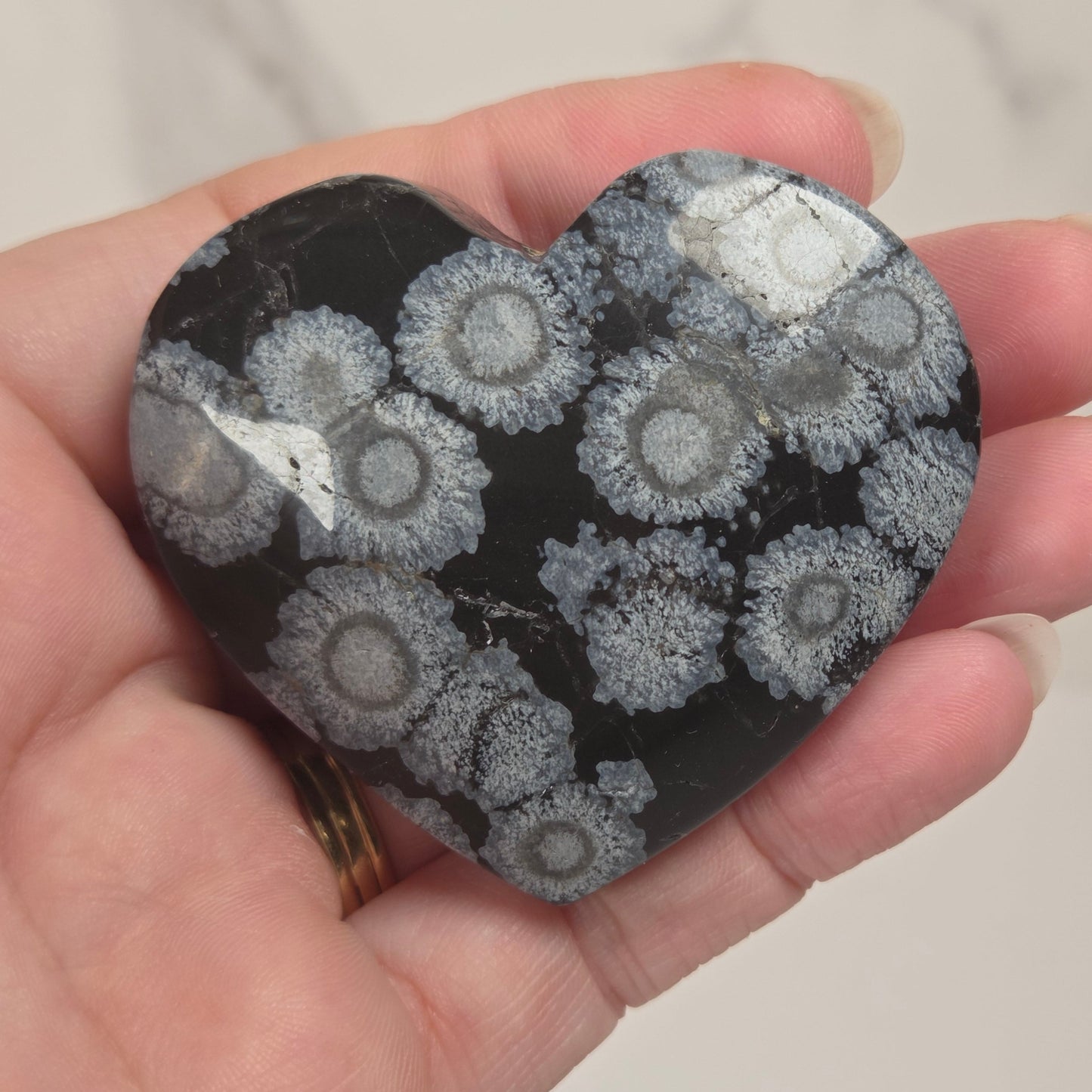 A heart-shaped polished stone featuring a black background with white and gray coral or floral concentric patterns, held in a person's hand against a light marble-look wall.