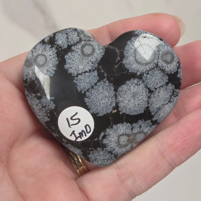 A heart-shaped polished stone with a black and gray coral-like pattern, held in a person's hand. A small white sticker on the stone reads '15 JND'.