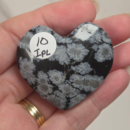 A heart-shaped polished stone with a black and gray mottled pattern, held in a person's hand. A small white circular sticker with handwritten text '10 IPL' is attached to the stone.
