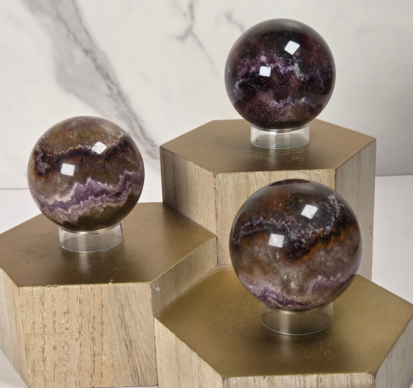 Three polished purple and brown agate crystal spheres displayed on hexagonal wooden pedestals with metallic gold caps, set against a white marble background.