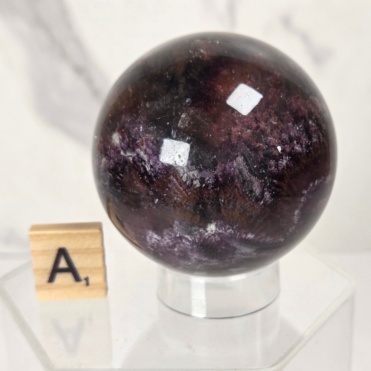 A polished dark purple crystal sphere with natural marbled patterns, displayed on a clear acrylic stand next to a wooden Scrabble tile showing the letter 'A'.