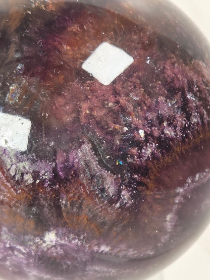 Close-up of a polished gemstone with a rounded shape, showing a mottled pattern of deep purple, burgundy, and brown with white specular highlights.