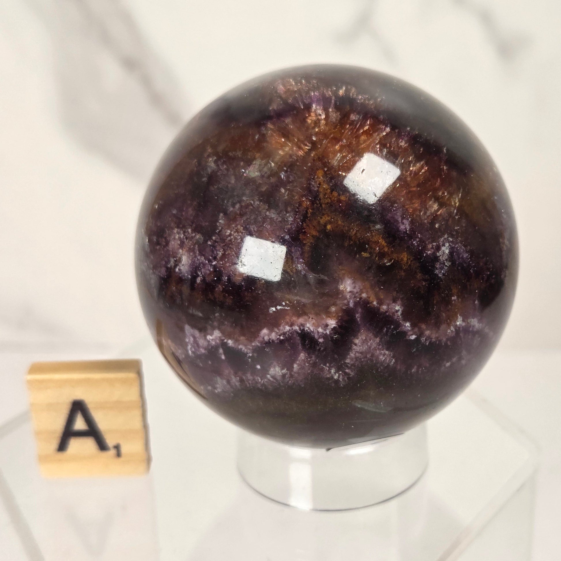 A polished dark purple and brown crystal sphere with metallic coppery highlights, displayed on a clear acrylic stand next to a wooden Scrabble tile showing the letter 'A'.