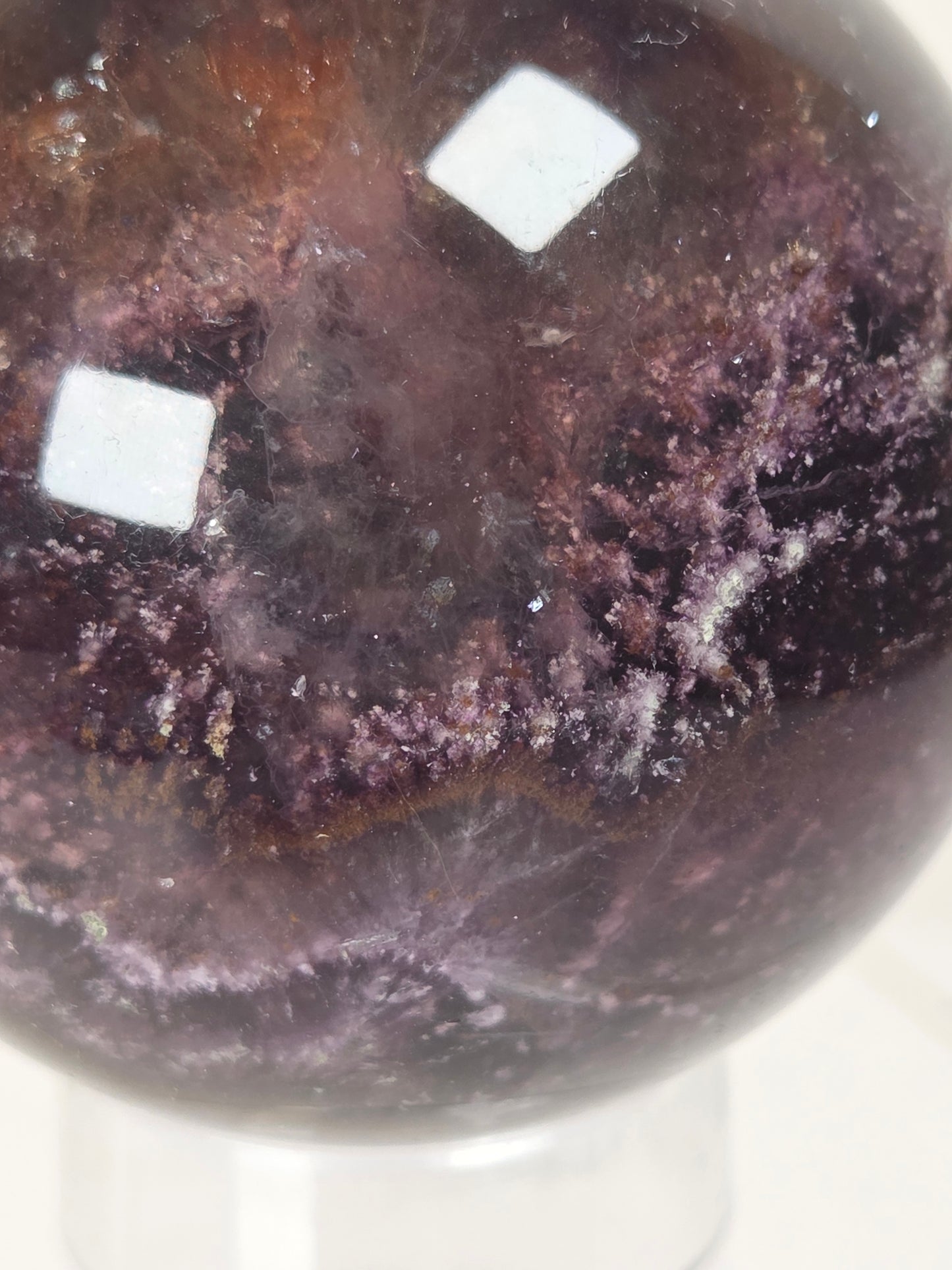 A close-up of a polished dark purple crystal sphere with natural mineral textures and two white square stickers on the surface, displayed on a clear stand against a light background.