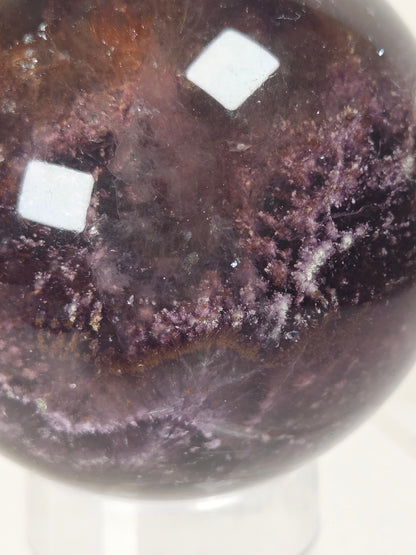 A close-up of a polished dark purple crystal sphere with natural mineral textures and two white square stickers on the surface, displayed on a clear stand against a light background.