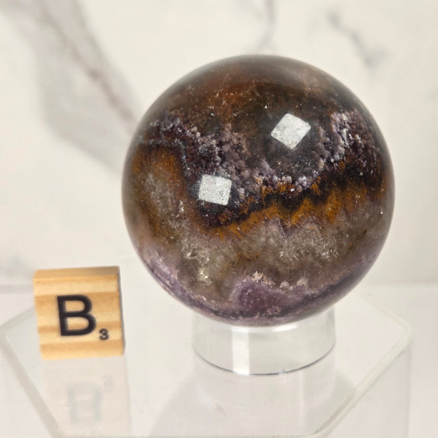 A polished agate crystal sphere with a marbled pattern of brown, purple, and gray, displayed on a clear acrylic stand next to a wooden Scrabble tile on a white marble surface.