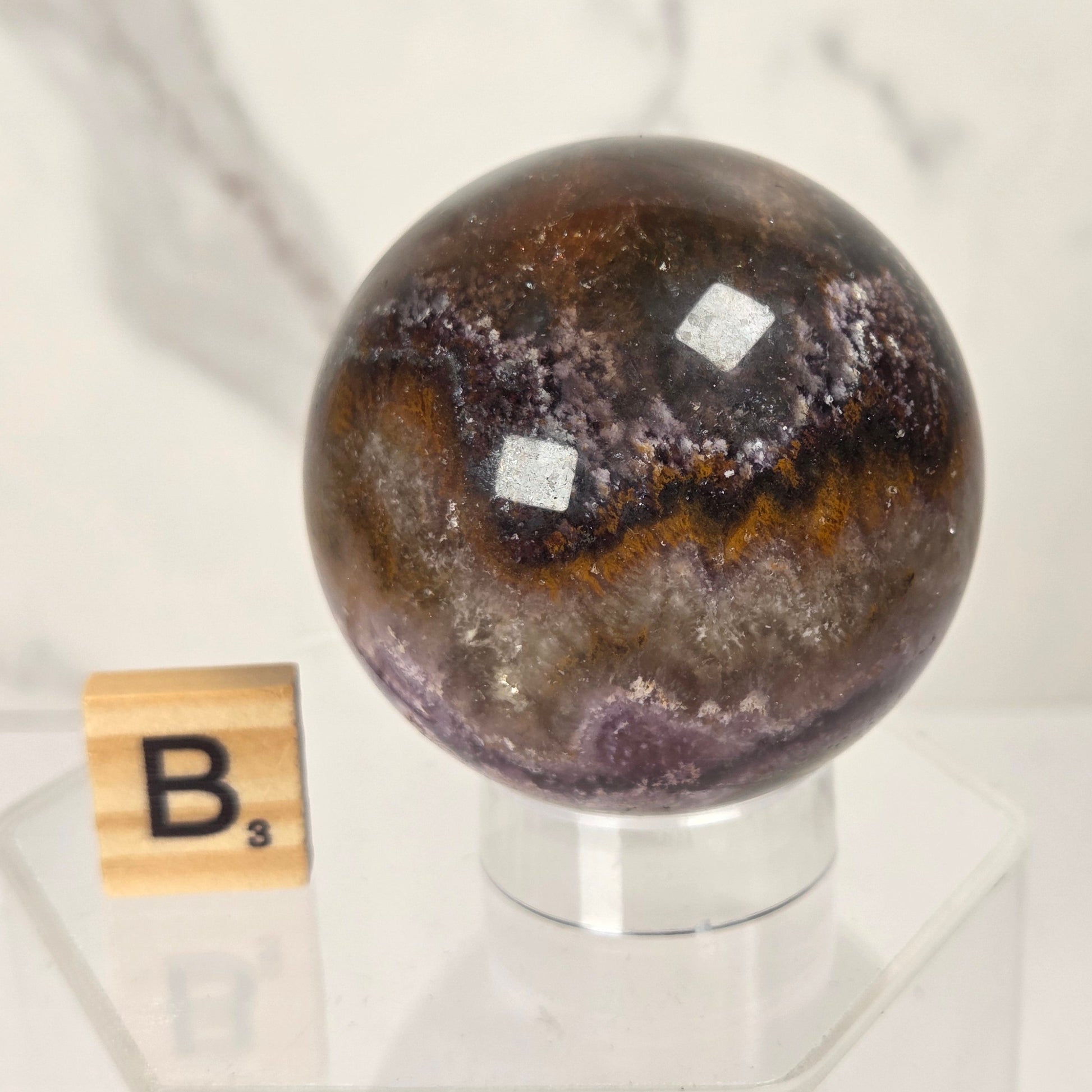 A polished agate crystal sphere with a marbled pattern of brown, purple, and gray, displayed on a clear acrylic stand next to a wooden Scrabble tile on a white marble surface.