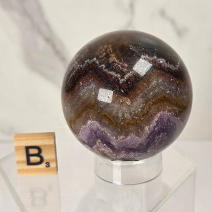 A polished agate crystal sphere with brown, black, and purple banding patterns, displayed on a clear acrylic stand next to a wooden Scrabble tile on a white marble surface.