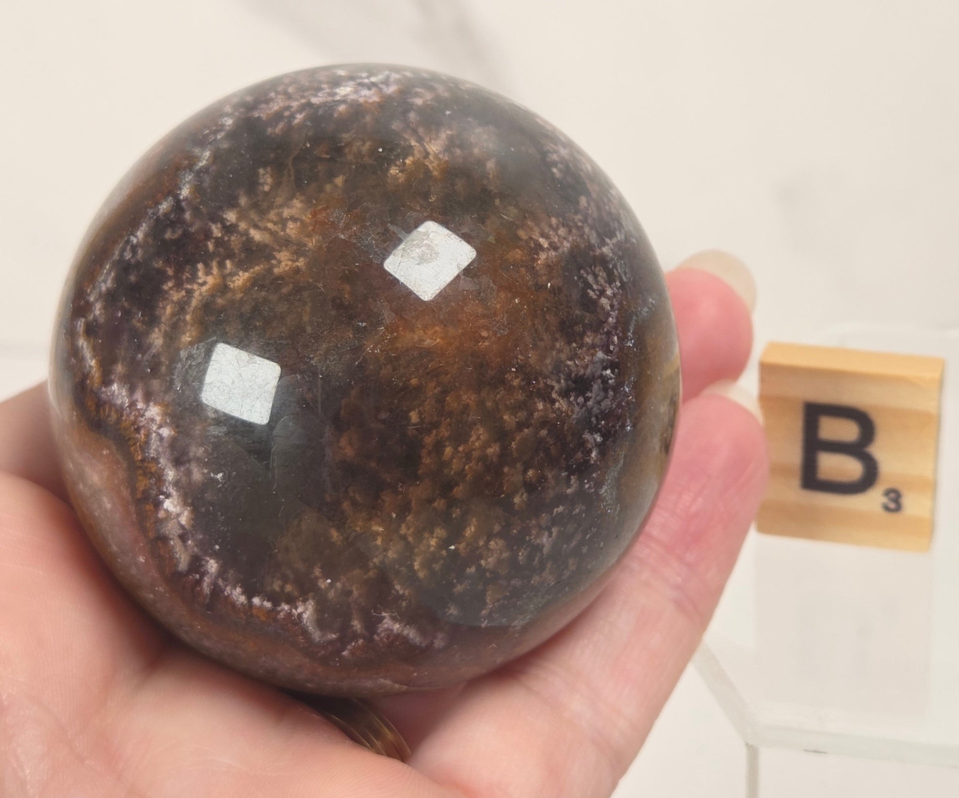 A hand holds a polished dark brown and black stone sphere with a mottled, metallic-like texture. A small wooden Scrabble tile showing the letter 'B' is visible in the background against a light marble-patterned wall.