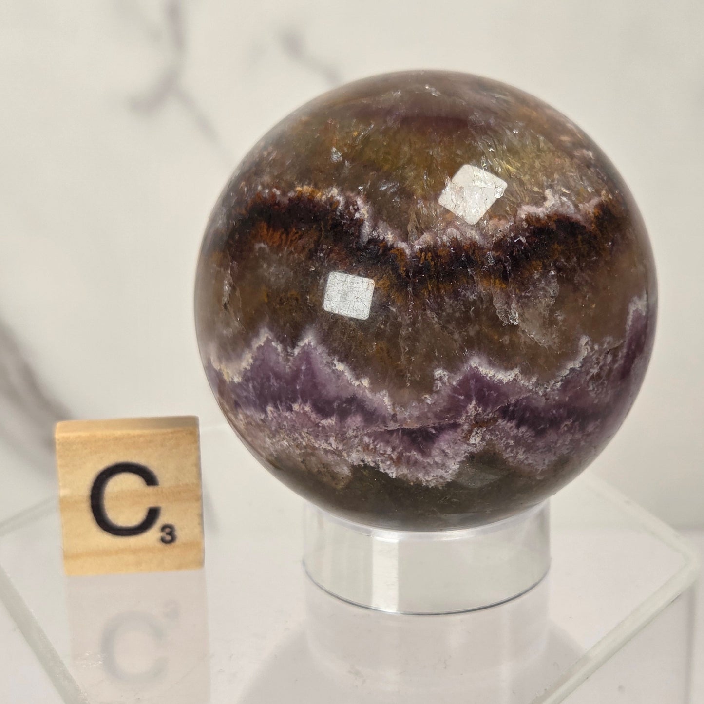 A polished crystal sphere with concentric bands of brown, purple, and white mineral patterns, displayed on a clear acrylic stand next to a Scrabble tile showing the letter C3.