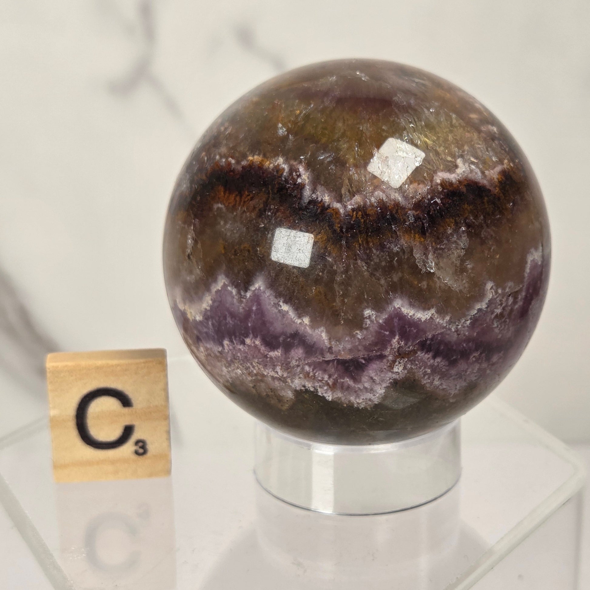 A polished crystal sphere with concentric bands of brown, purple, and white mineral patterns, displayed on a clear acrylic stand next to a Scrabble tile showing the letter C3.