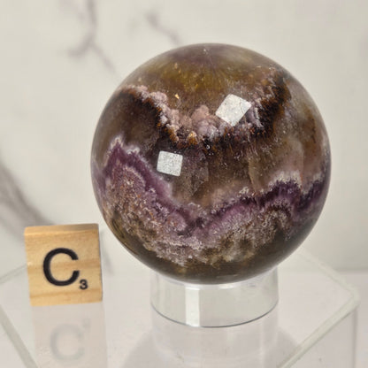 A polished crystal sphere with concentric bands of brown, purple, and white mineral patterns, displayed on a clear acrylic stand next to a small wooden Scrabble tile showing the letter 'C'.