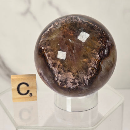 A polished brown and amber glass orb with two small white labels, displayed on a clear acrylic stand next to a wooden Scrabble tile showing the letter C.