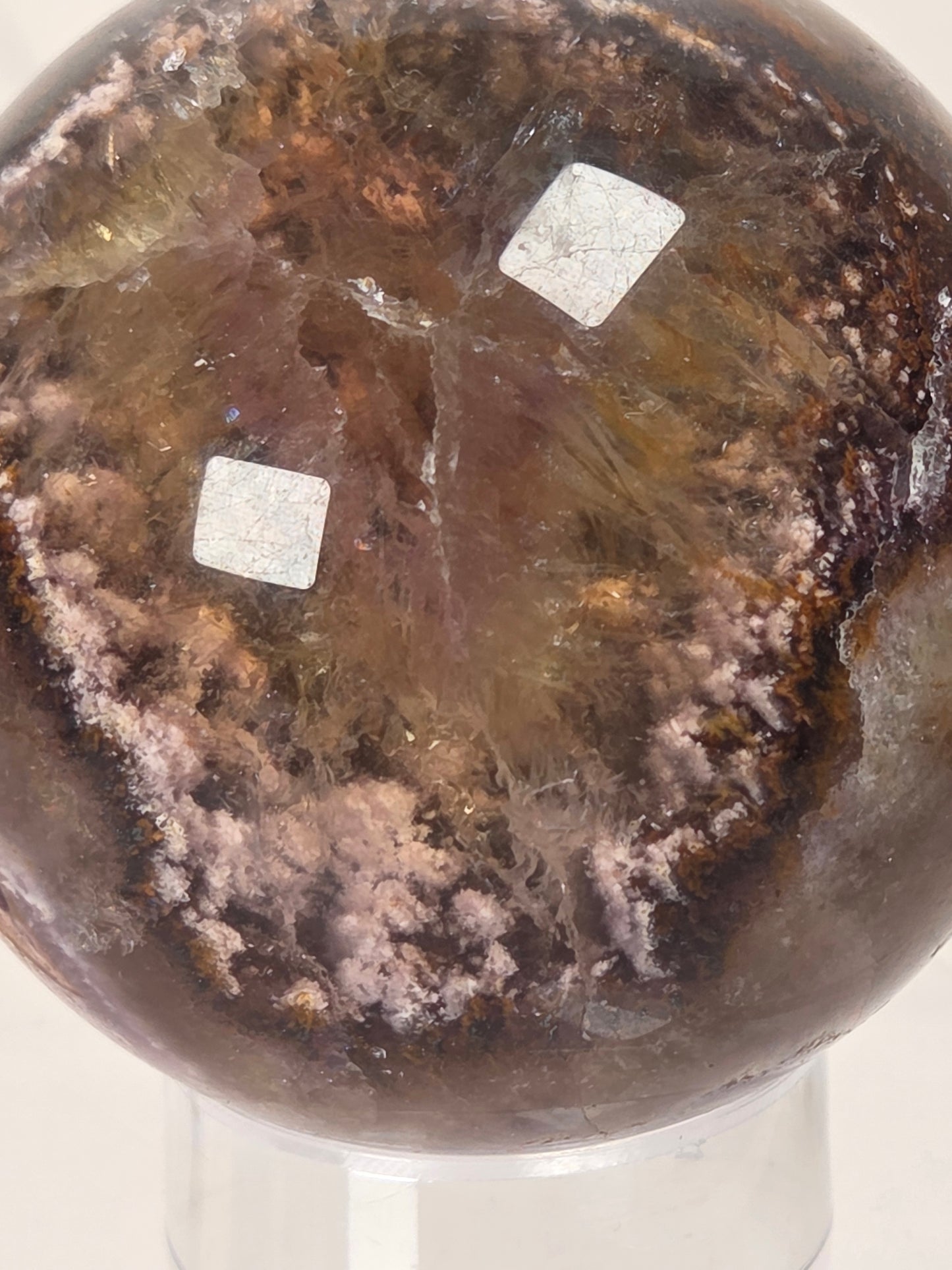 A polished brown crystal sphere with pinkish-purple and amber inclusions, displayed on a clear stand against a light background.