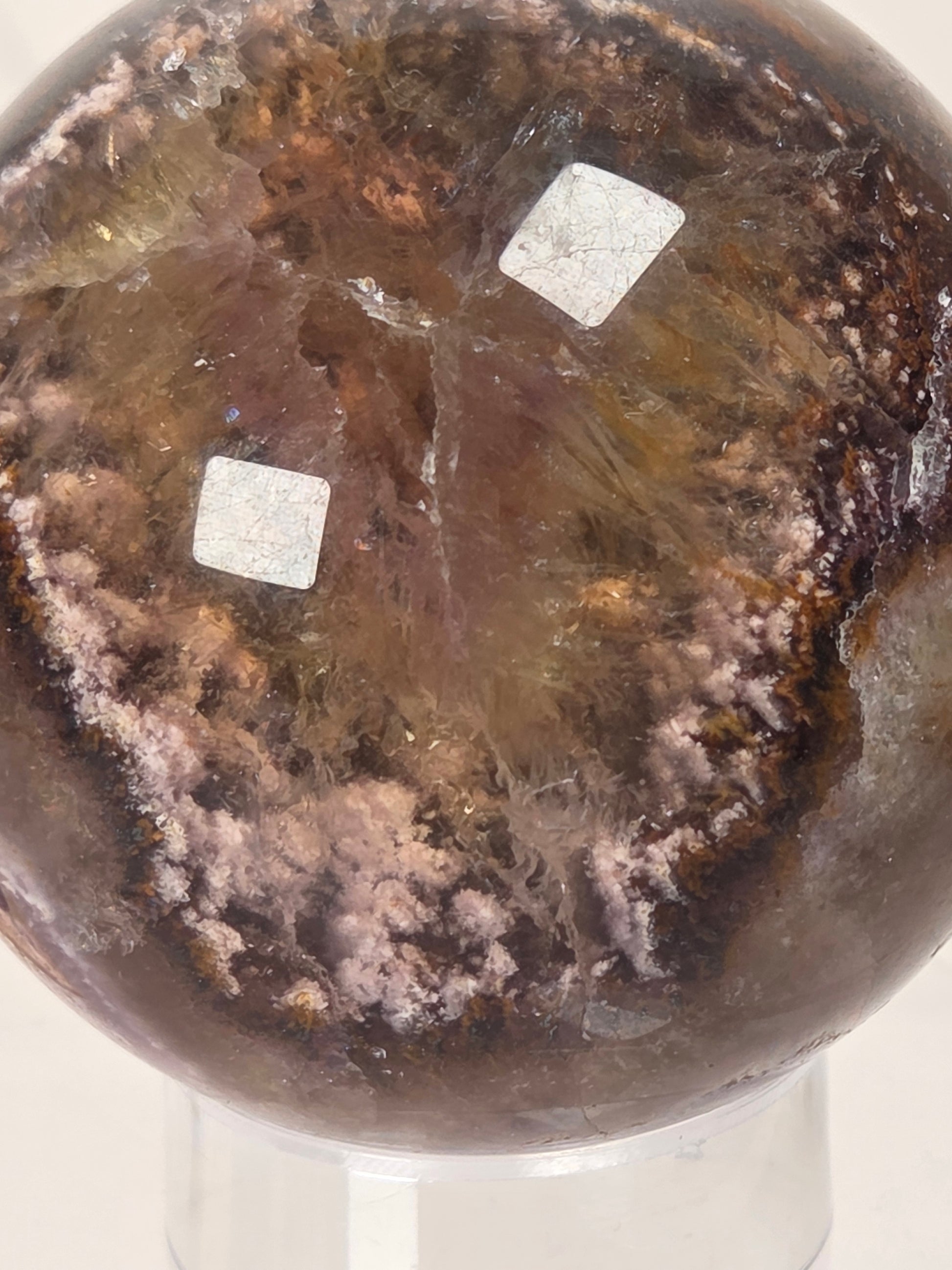 A polished brown crystal sphere with pinkish-purple and amber inclusions, displayed on a clear stand against a light background.