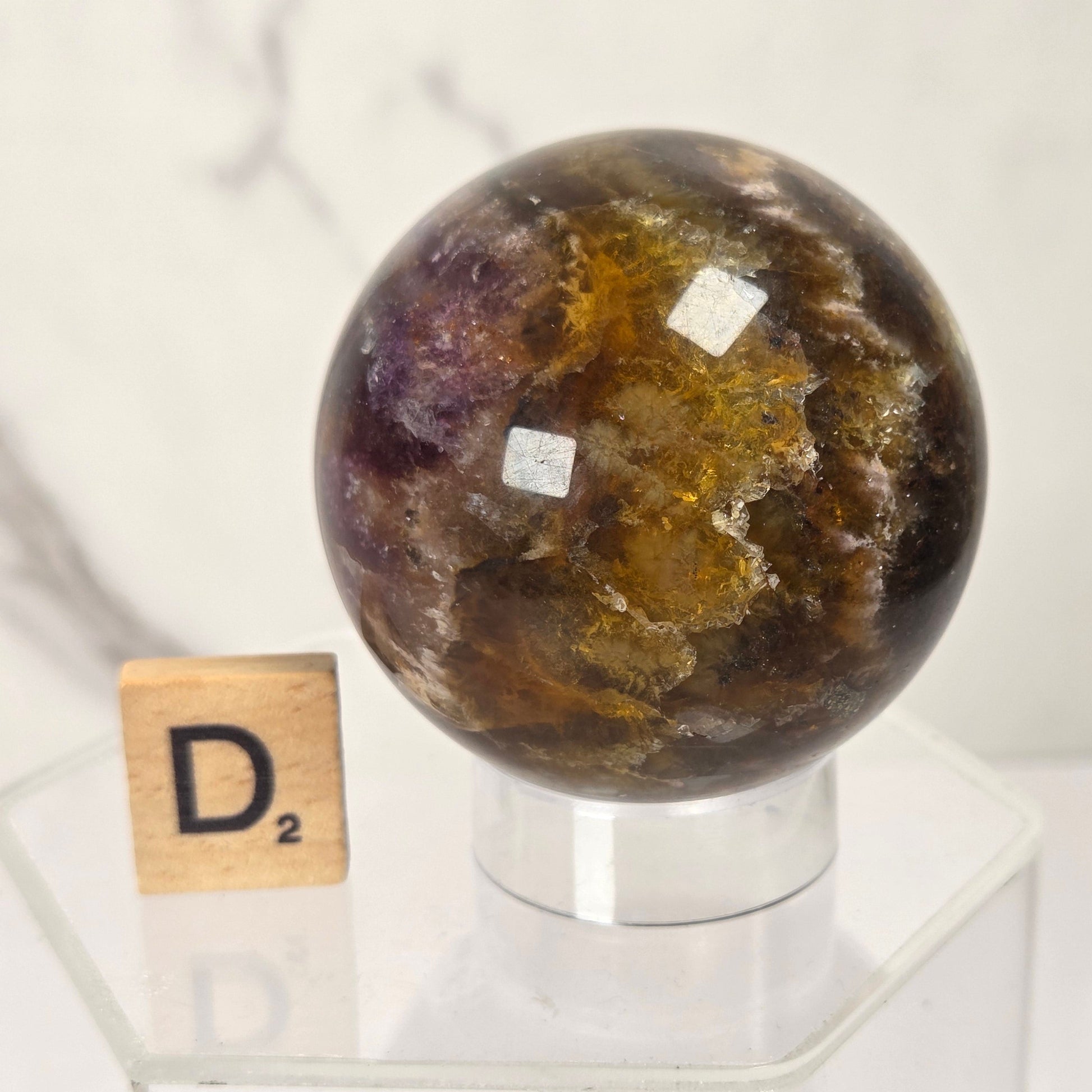 A polished crystal sphere with mottled brown, amber, and purple tones displayed on a clear acrylic stand next to a Scrabble tile showing the letter D.