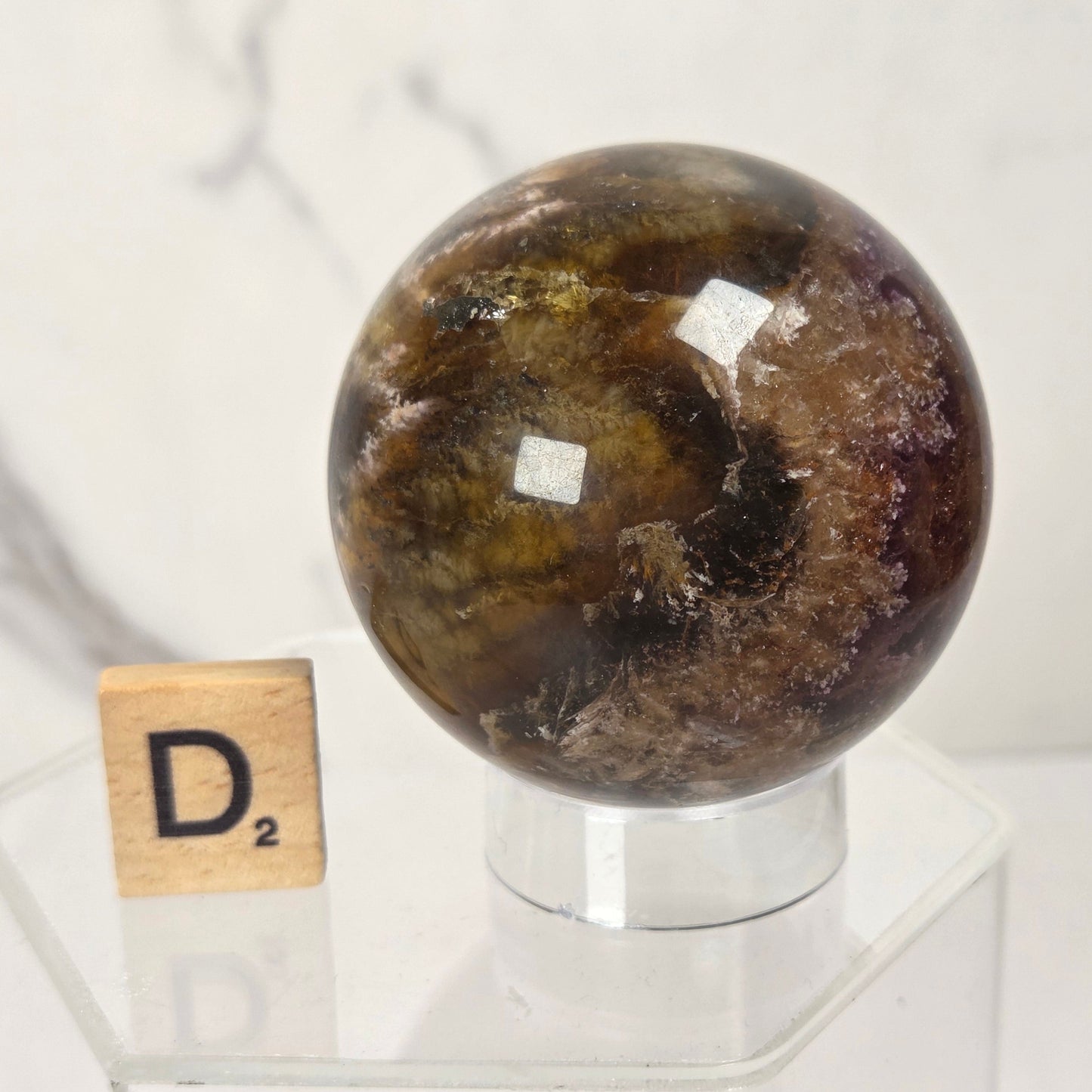 A polished brown and gold jasper sphere on a clear acrylic display stand, with a small wooden Scrabble tile showing the letter D2 placed beside it on a light marble surface.