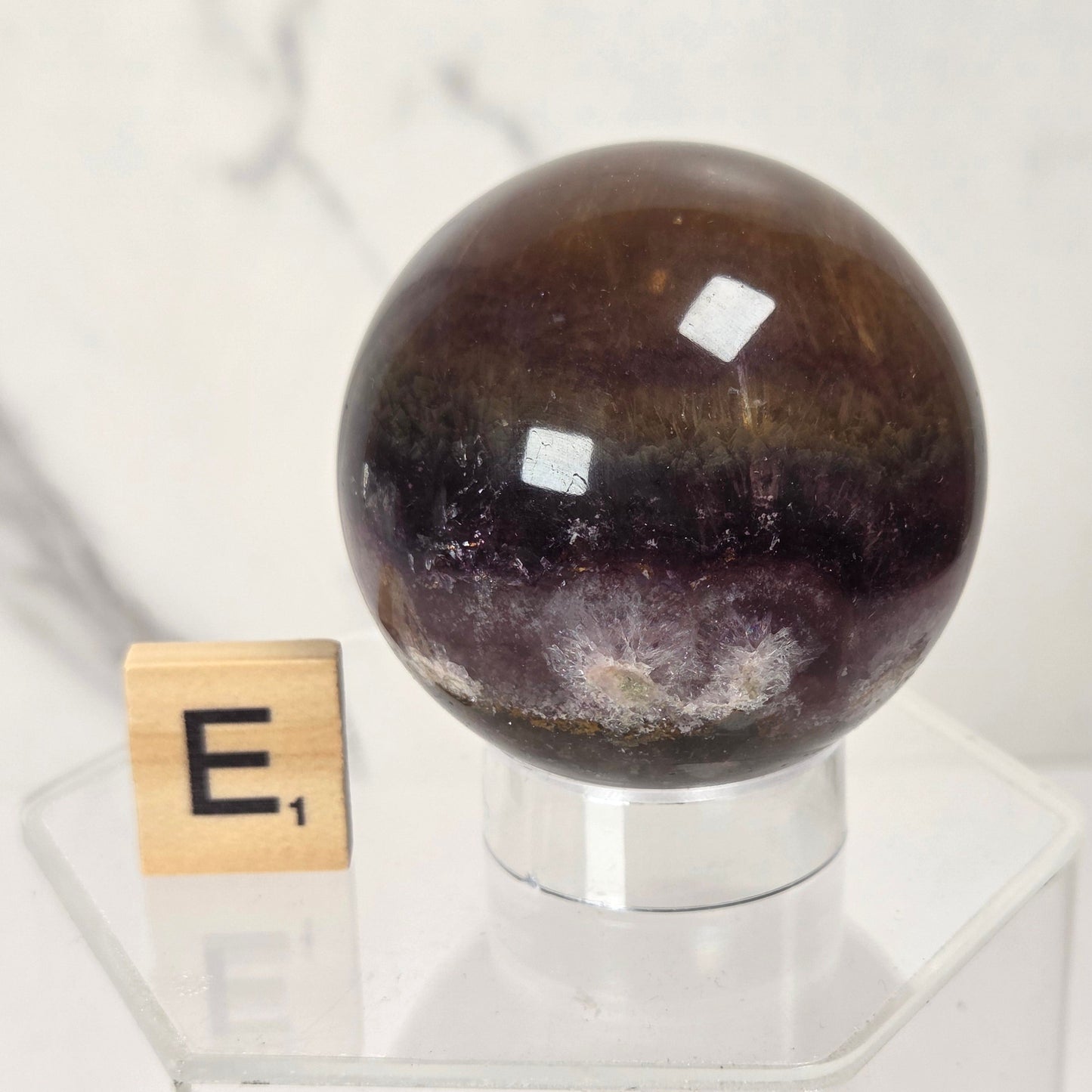 A polished dark brown and purple amethyst crystal sphere displayed on a clear acrylic stand next to a wooden Scrabble tile with the letter 'E', set against a light marble background.