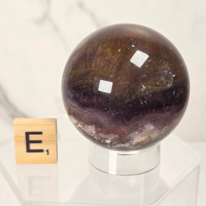 A polished dark brown and purple agate crystal sphere displayed on a clear acrylic stand next to a small wooden Scrabble tile with the letter 'E', set against a light marble background.