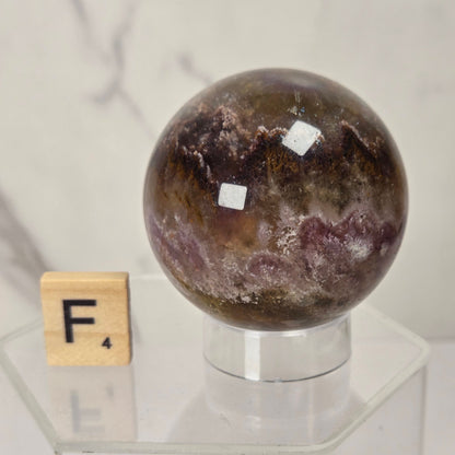 A polished crystal sphere with mottled brown, purple, and cream patterns, displayed on a clear acrylic stand next to a wooden Scrabble tile showing the letter F.