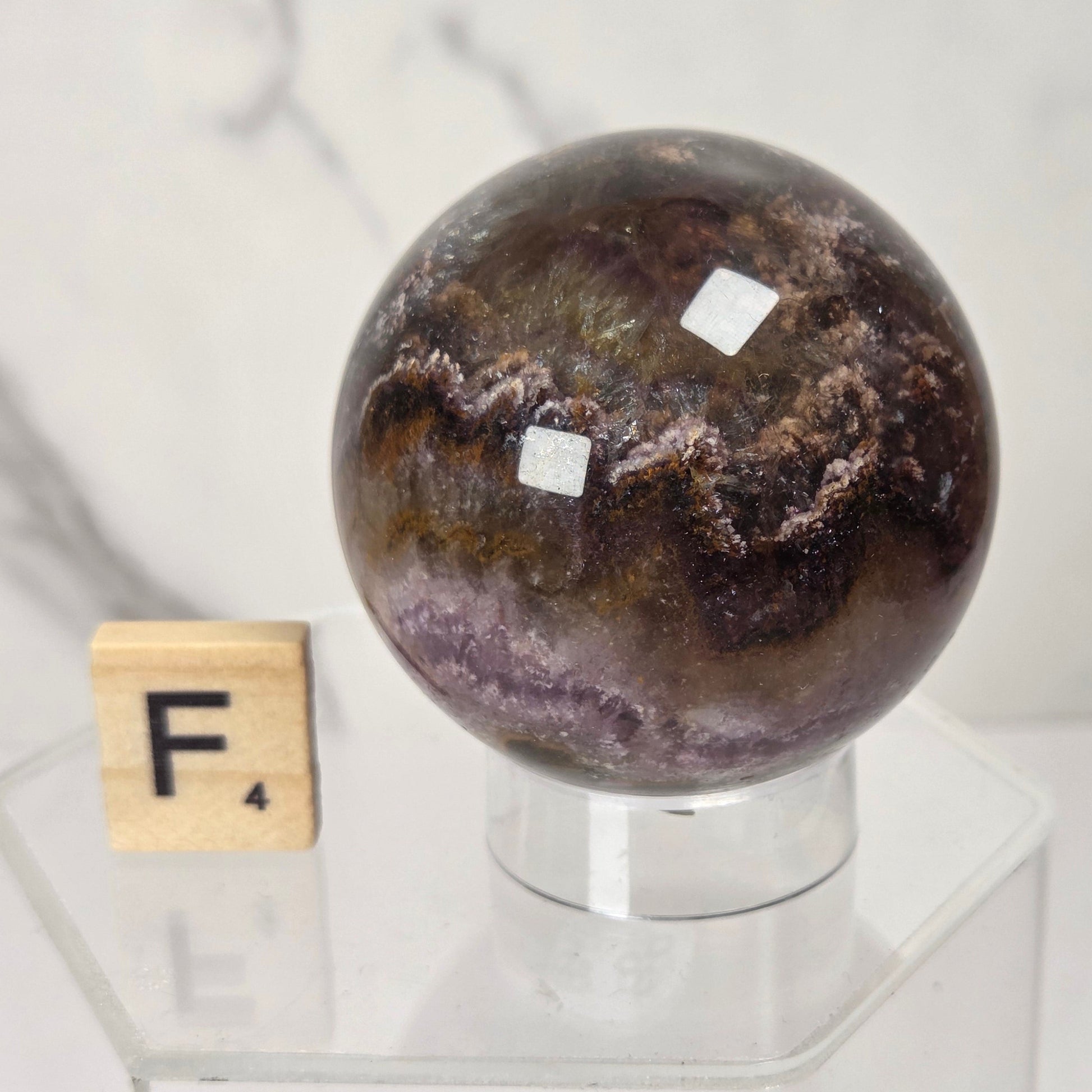 A polished dark brown and purple agate crystal sphere displayed on a clear acrylic stand next to a wooden Scrabble tile with the letter F, set against a light marble background.