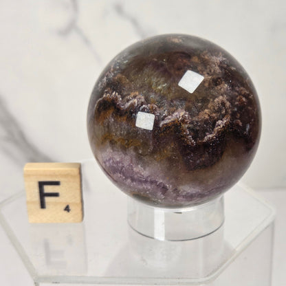 A polished dark brown and purple agate crystal sphere displayed on a clear acrylic stand next to a wooden Scrabble tile with the letter F, set against a light marble background.