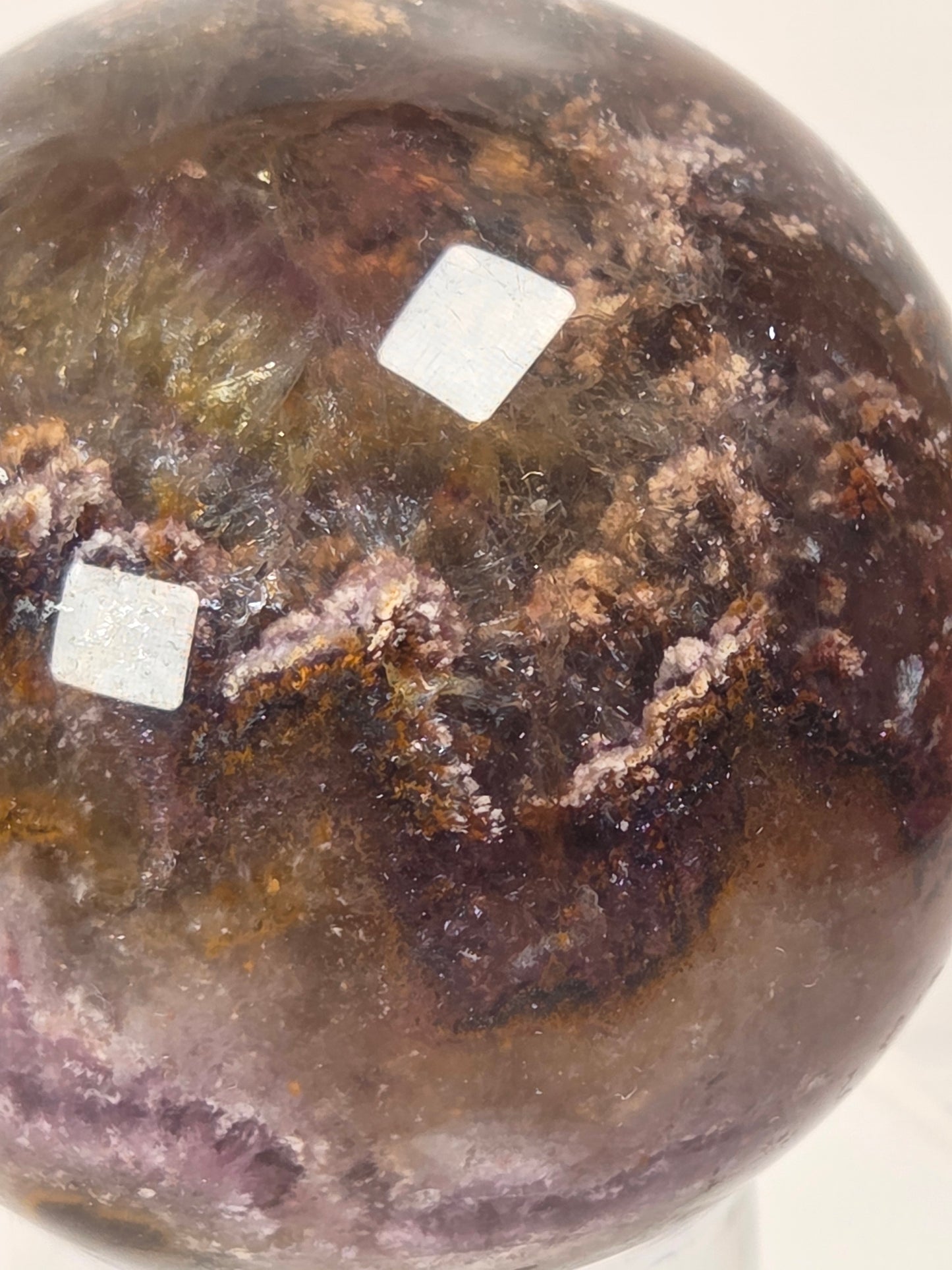 A close-up of a polished round crystal sphere with natural mineral inclusions in shades of brown, purple, and amber, with two small white square stickers on the surface.