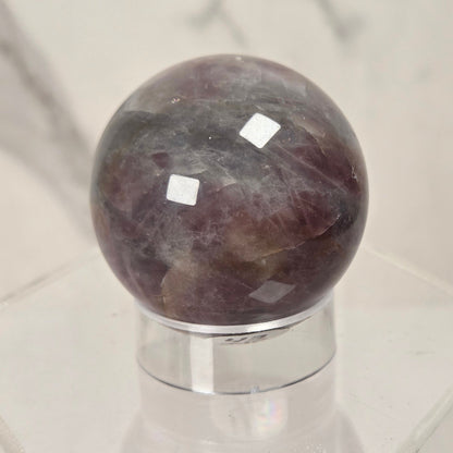 A polished round crystal sphere with a marbled purple, gray, and white pattern, displayed on a clear acrylic stand against a light marble-patterned background.