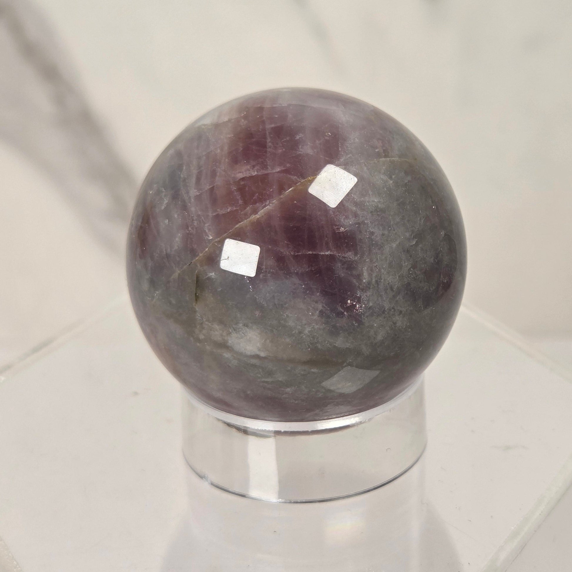 A polished purple and gray stone sphere displayed on a clear acrylic stand against a light marble-patterned background.