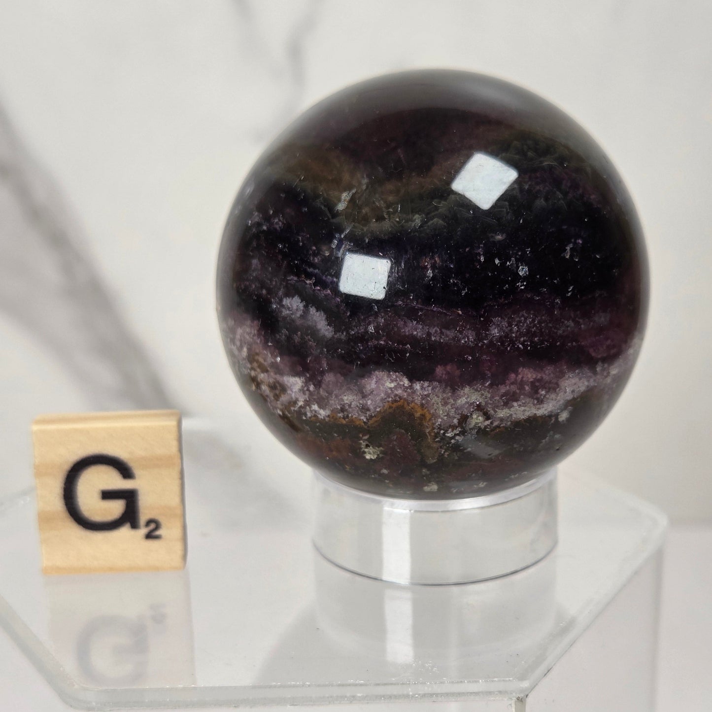 A polished dark purple and black agate crystal sphere displayed on a clear acrylic stand next to a wooden Scrabble tile showing the letter G.