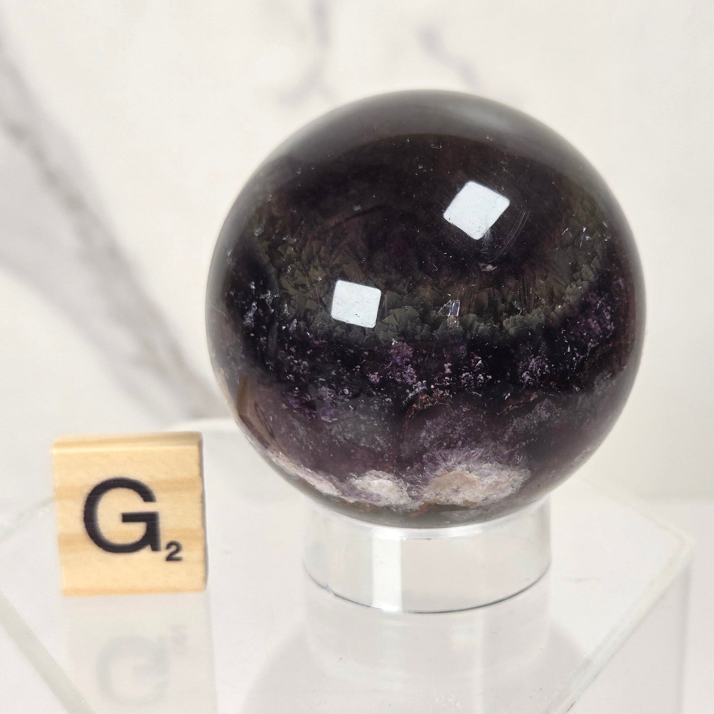 A polished dark purple crystal sphere on a clear acrylic display stand, with a small wooden letter tile showing 'G2' beside it, set against a light marble-patterned background.