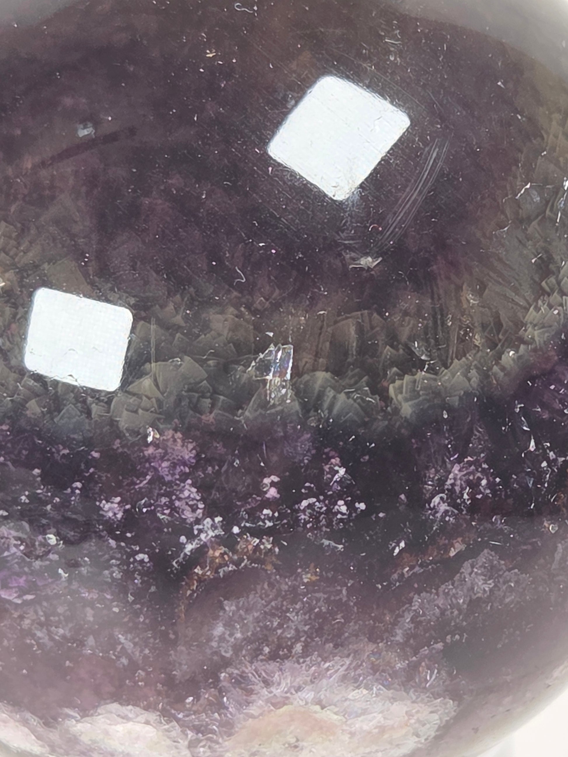 A close-up of a polished crystal sphere, likely amethyst, showing deep purple and violet tones with natural mineral inclusions and a smooth, rounded edge.