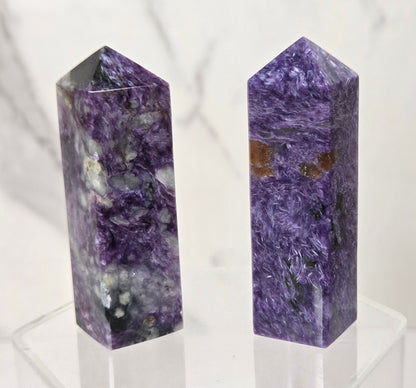 Two polished purple crystal chakra towers, one of Amethyst and one of Blue Sapphire, standing side-by-side on a clear acrylic display stand against a white marble background.
