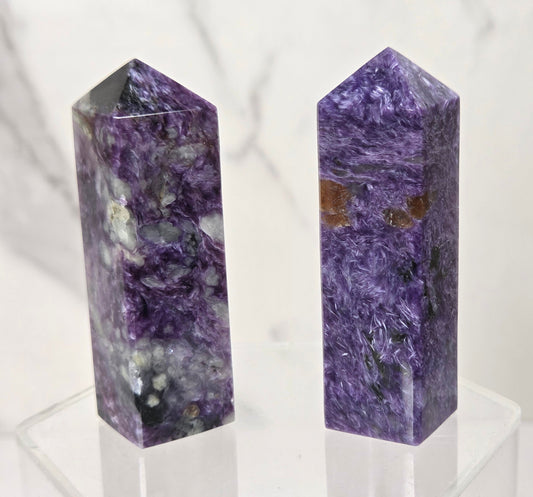 Two polished purple crystal chakra towers, one of Amethyst and one of Blue Sapphire, standing side-by-side on a clear acrylic display stand against a white marble background.