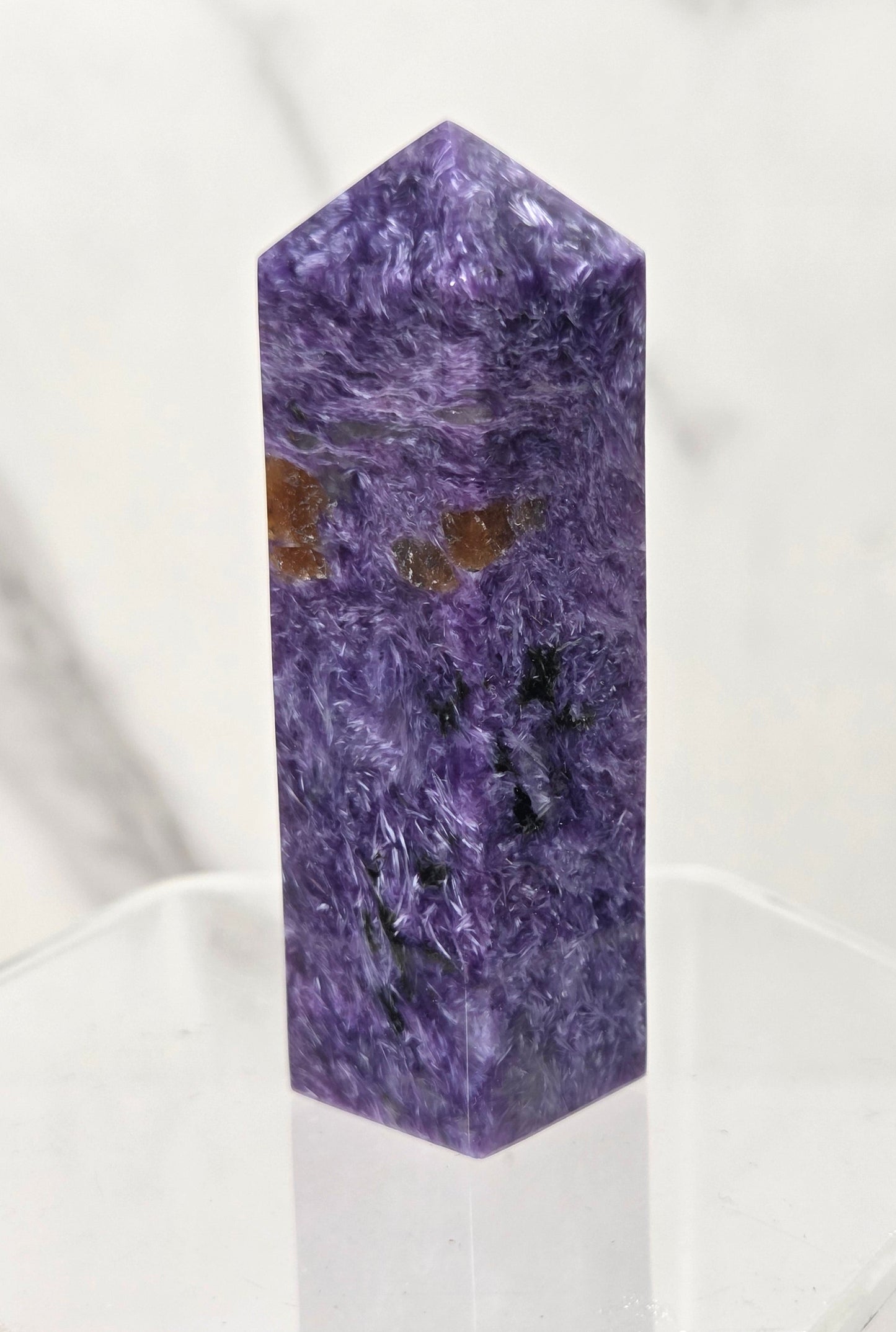 A polished purple crystal tower or point with natural marbling and small brown inclusions, displayed on a clear acrylic stand against a light marble-patterned background.