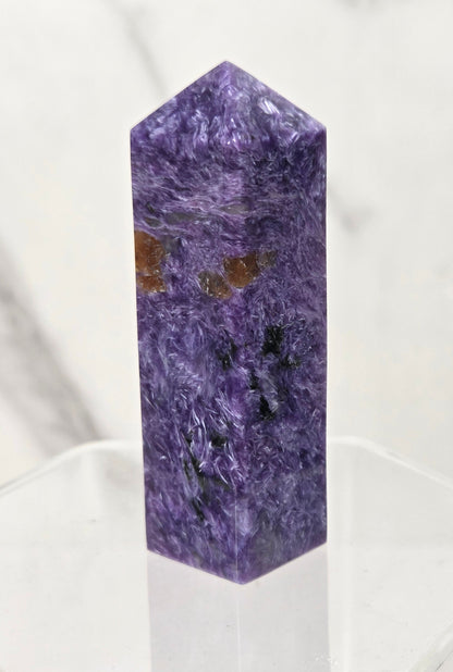 A polished purple crystal tower or point with natural marbling and small brown inclusions, displayed on a clear acrylic stand against a light marble-patterned background.