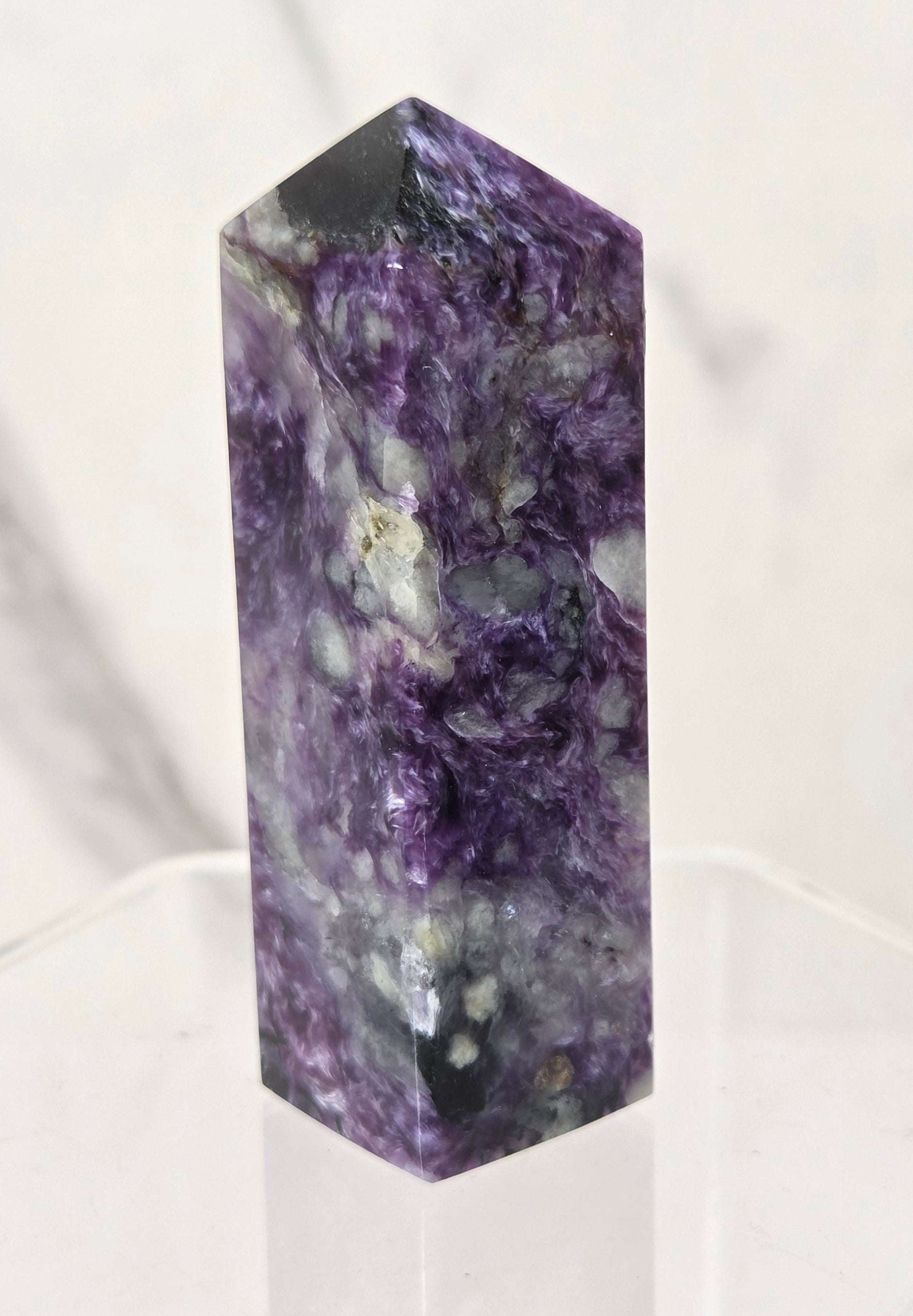 A polished purple fluorite crystal tower with natural marbling and white inclusions, standing upright on a clear acrylic hexagonal display base against a light marble-patterned background.