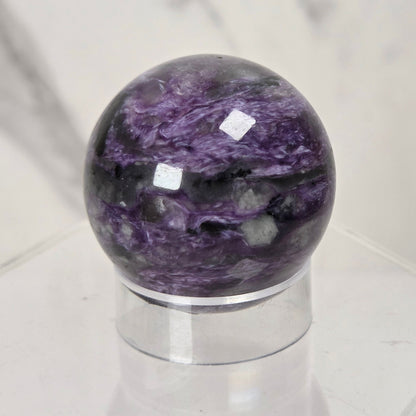 A polished purple and black marbled crystal sphere displayed on a clear acrylic stand against a light marble-patterned background.