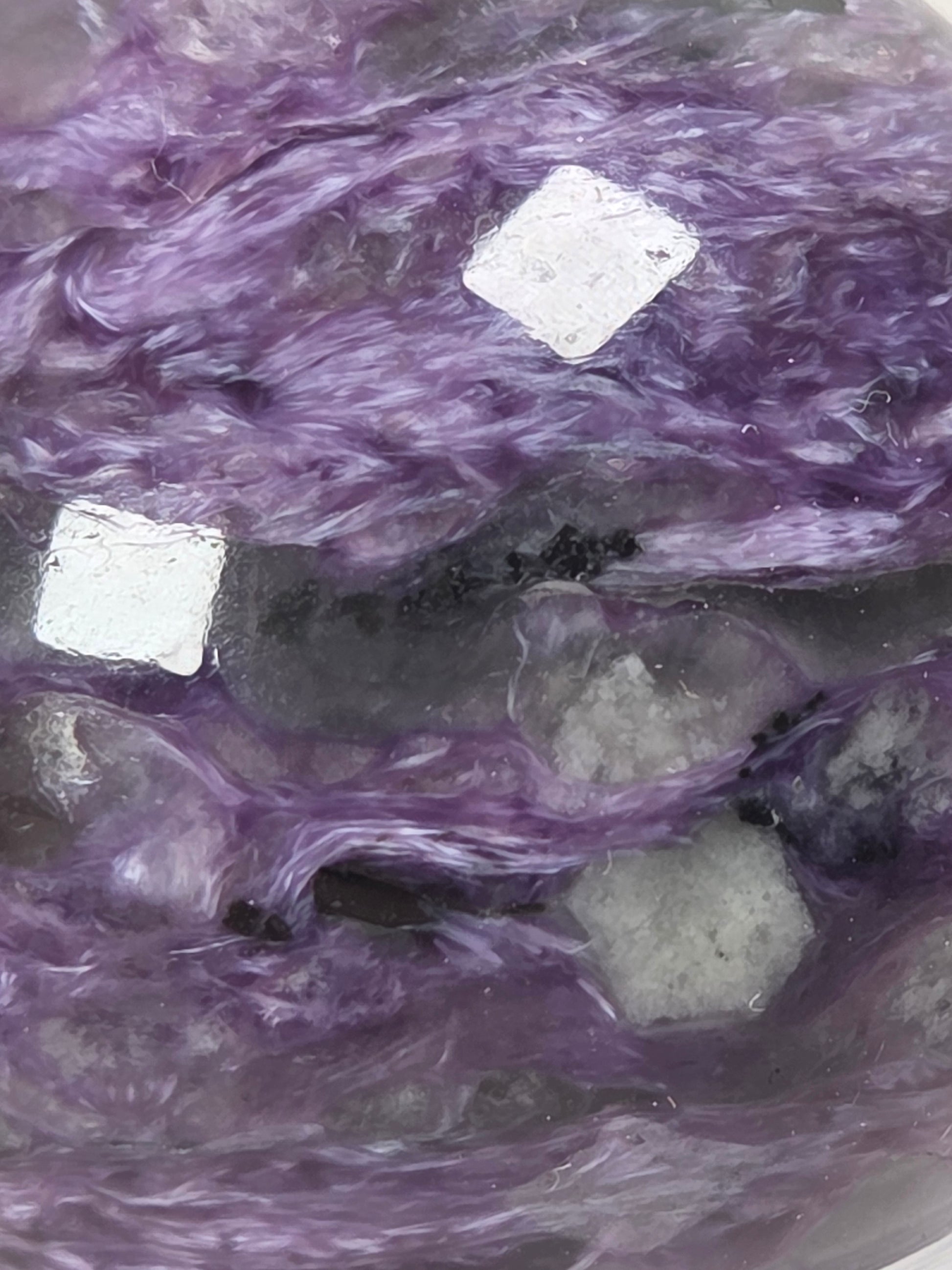 A close-up of a purple mineral specimen with layered fibrous texture, dark gray inclusions, and two small metallic silver chips placed on top.