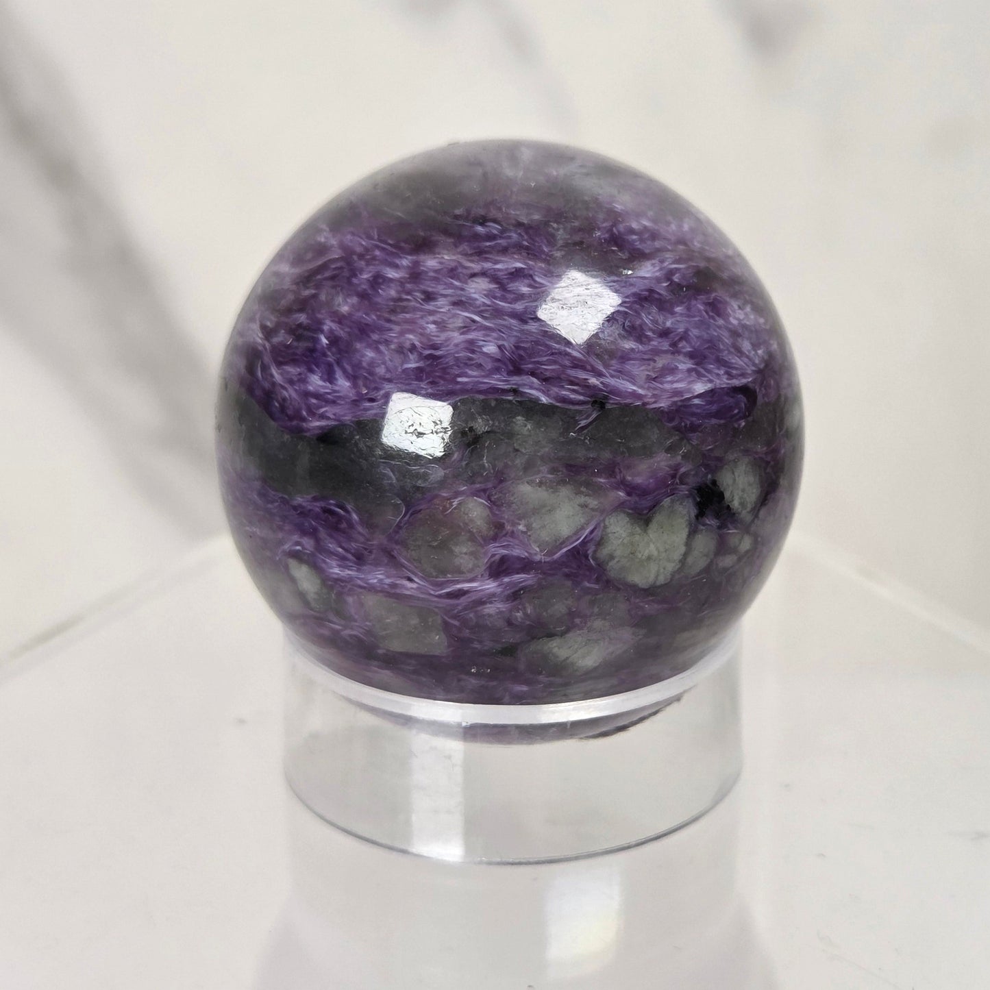A polished purple and gray marbled stone sphere displayed on a clear acrylic stand against a light marble-patterned background.