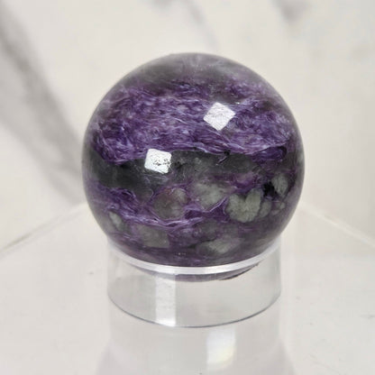 A polished purple and gray marbled stone sphere displayed on a clear acrylic stand against a light marble-patterned background.