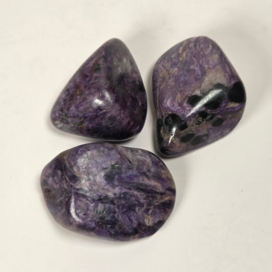 Three polished purple tumbled stones arranged on a plain off-white background.