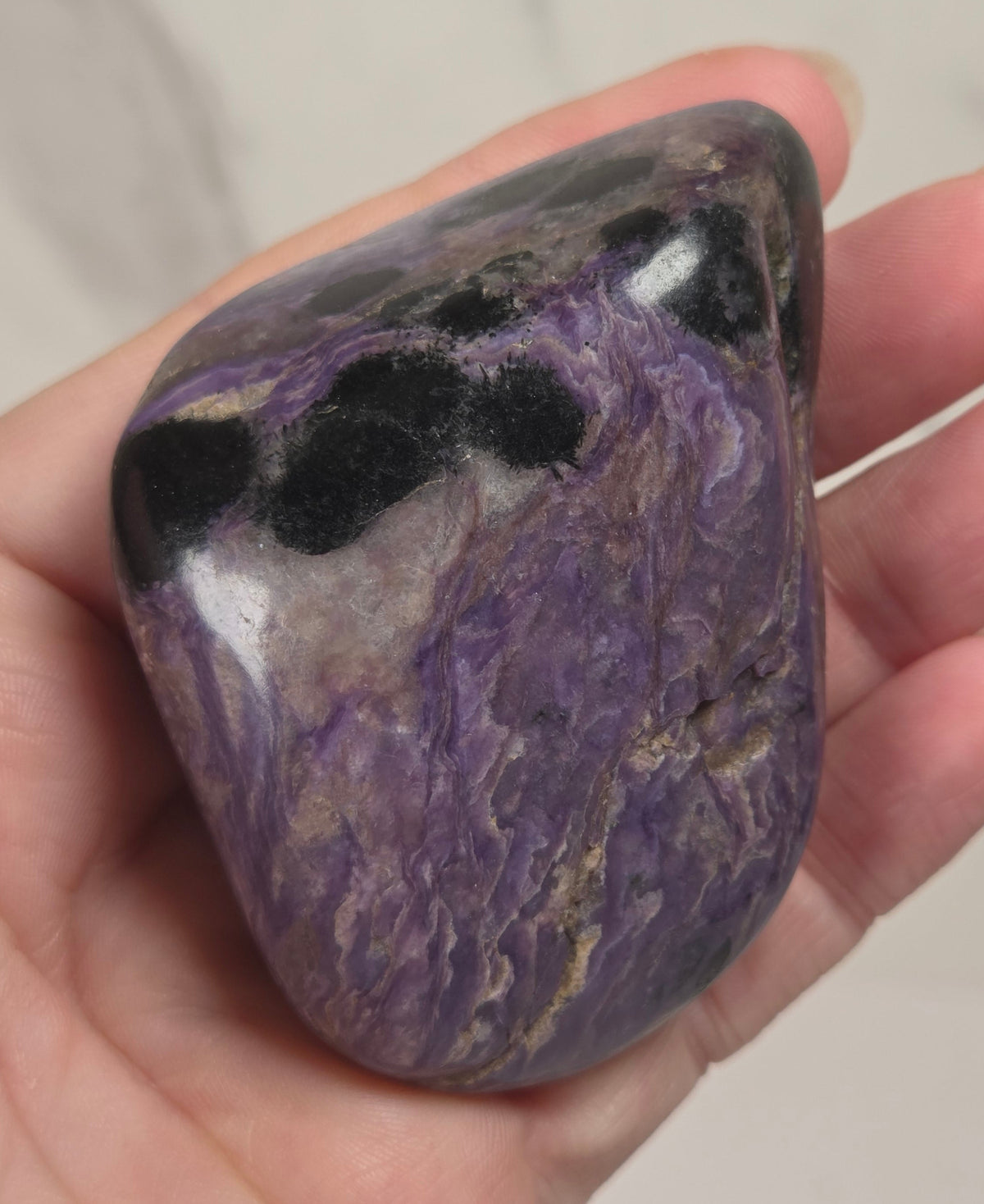 A hand holding a polished purple tumbled stone with black and white marbled patterns, set against a light marble background.