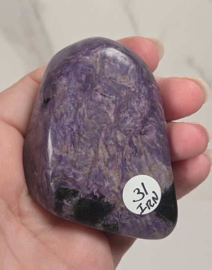 A hand holding a polished purple stone with natural marbled patterns and dark inclusions, featuring a small white price sticker on the side.